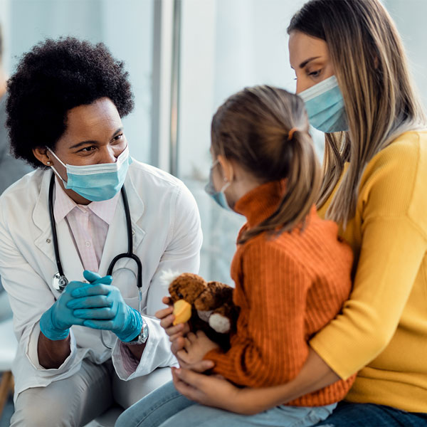 A doctor speaks with a young girl who is seated on her mother’s lap holding a stuffed toy.